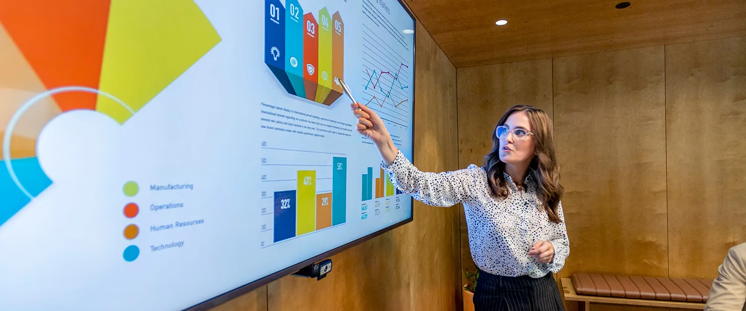 A woman gestures to a screen of data visualizations as she presents to a boardroom