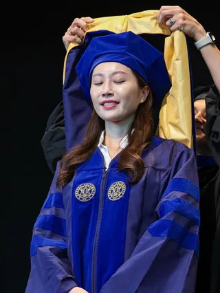 A PhD graduate smiles serenly while receiving their regalia on stage during commencement.